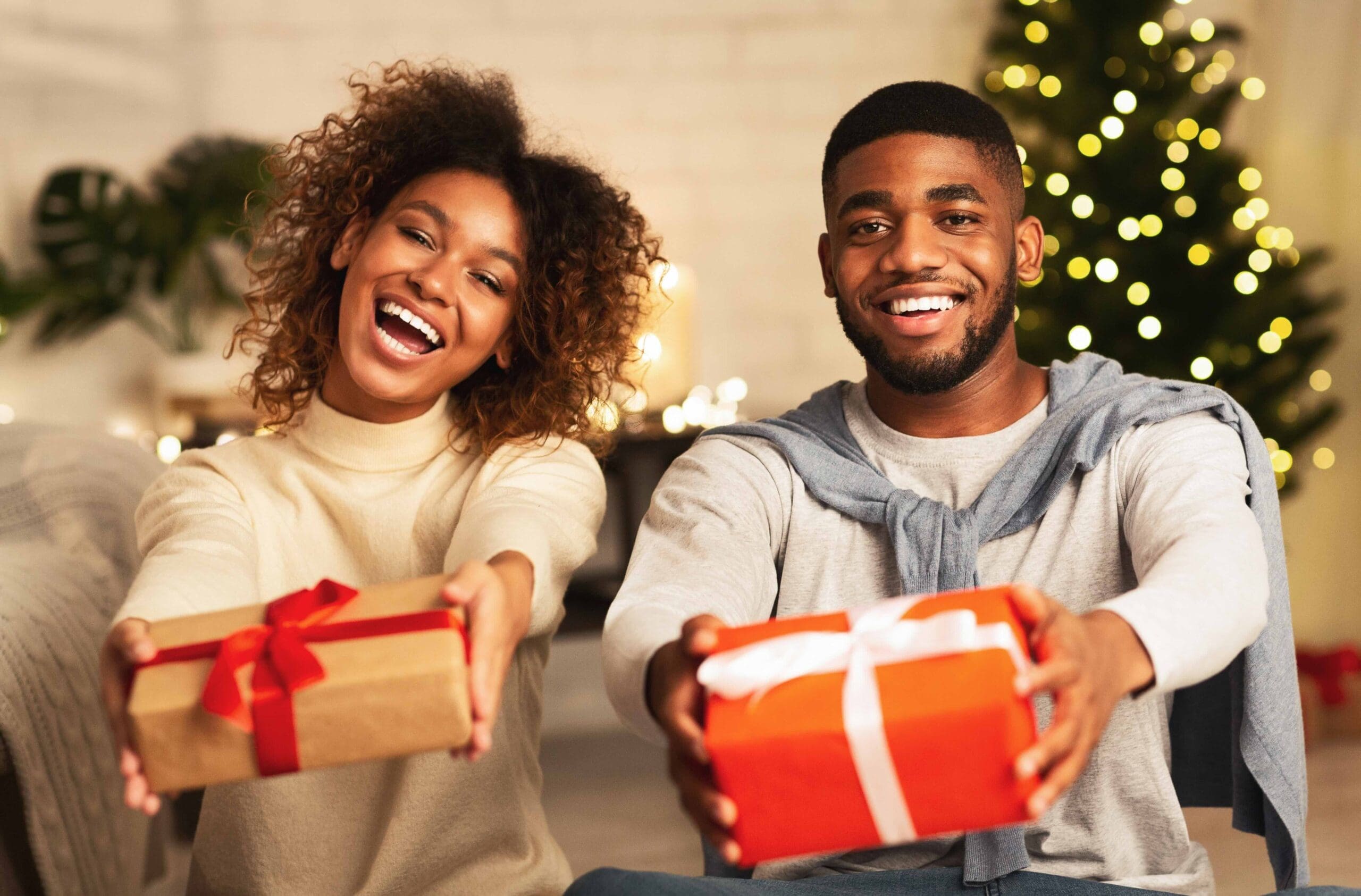 Black couple holding Christmas gift boxes and smiling in front of xmas tree