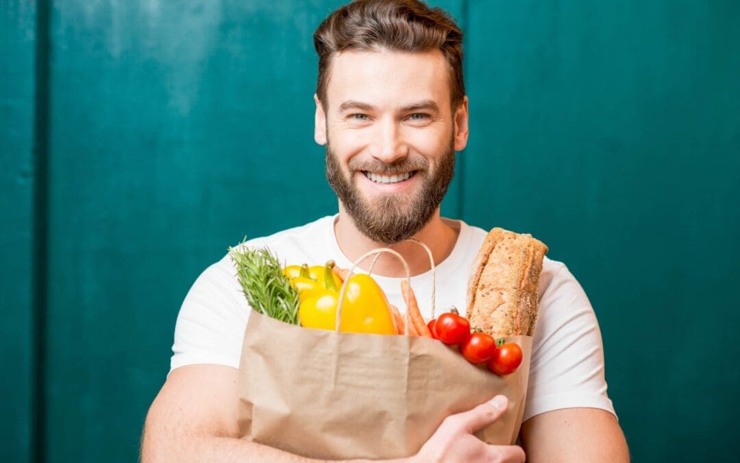 Handsome man with a white tshirt on holding a brown paper bag full of healthy food with a green background behind him.