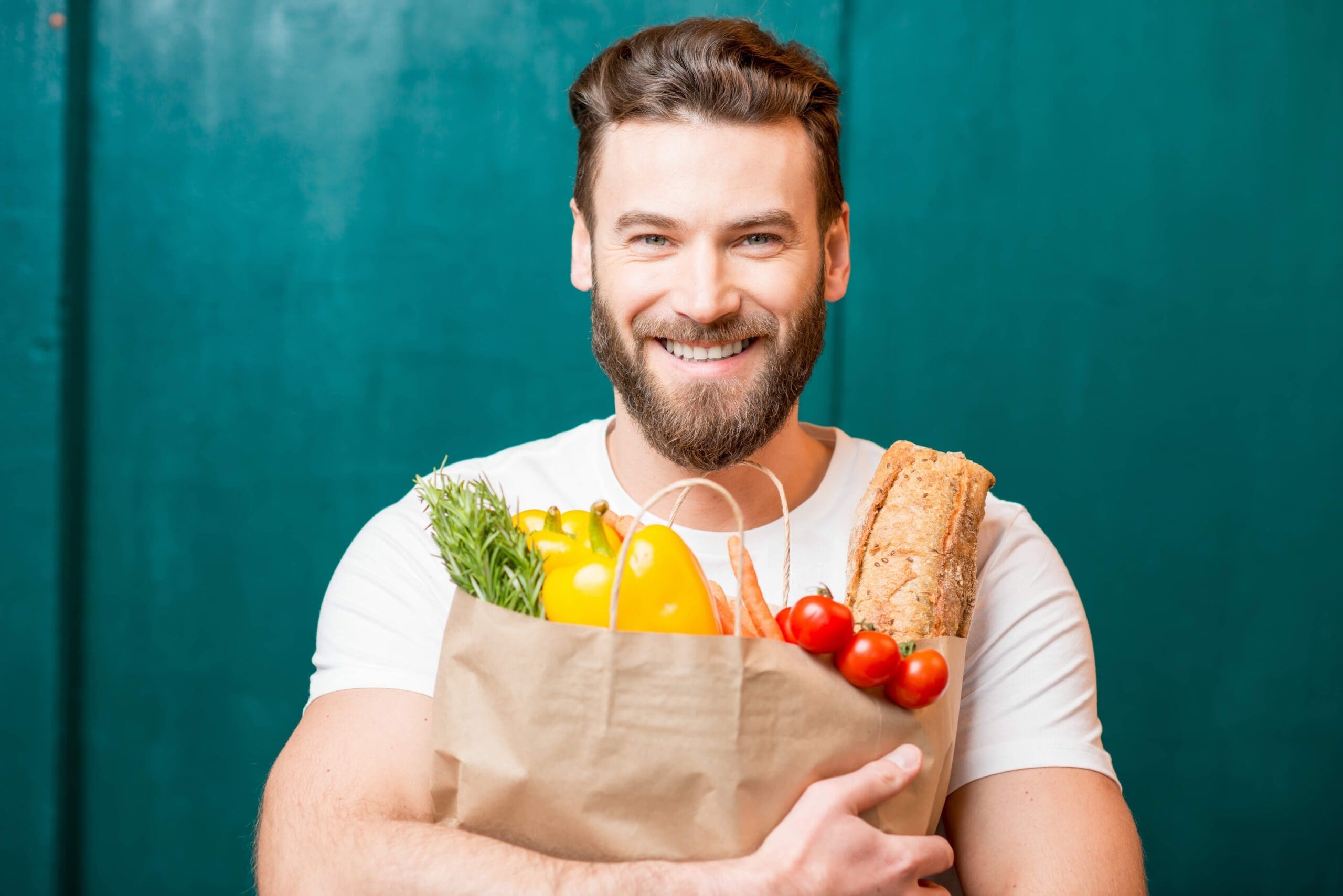 Beautiful happy girl in Santa Claus hat showing gift box Handsome man with a white tshirt on holding a brown paper bag full of healthy food with a green background behind him.