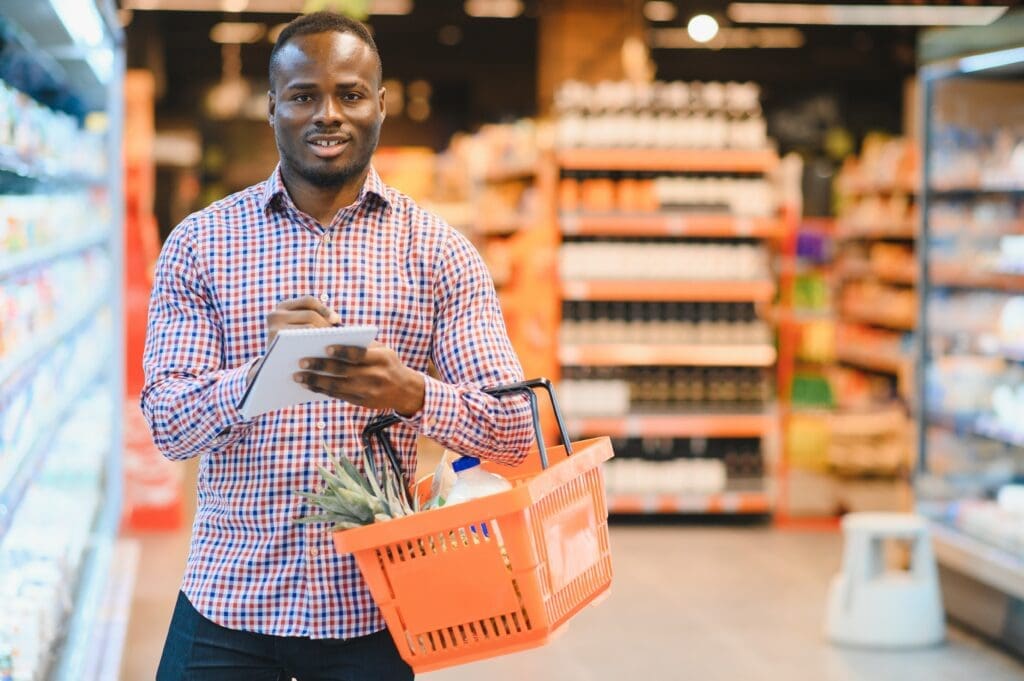 Black man wearing a plaid shirt shopping at supermarket holding orange shopping basket. 