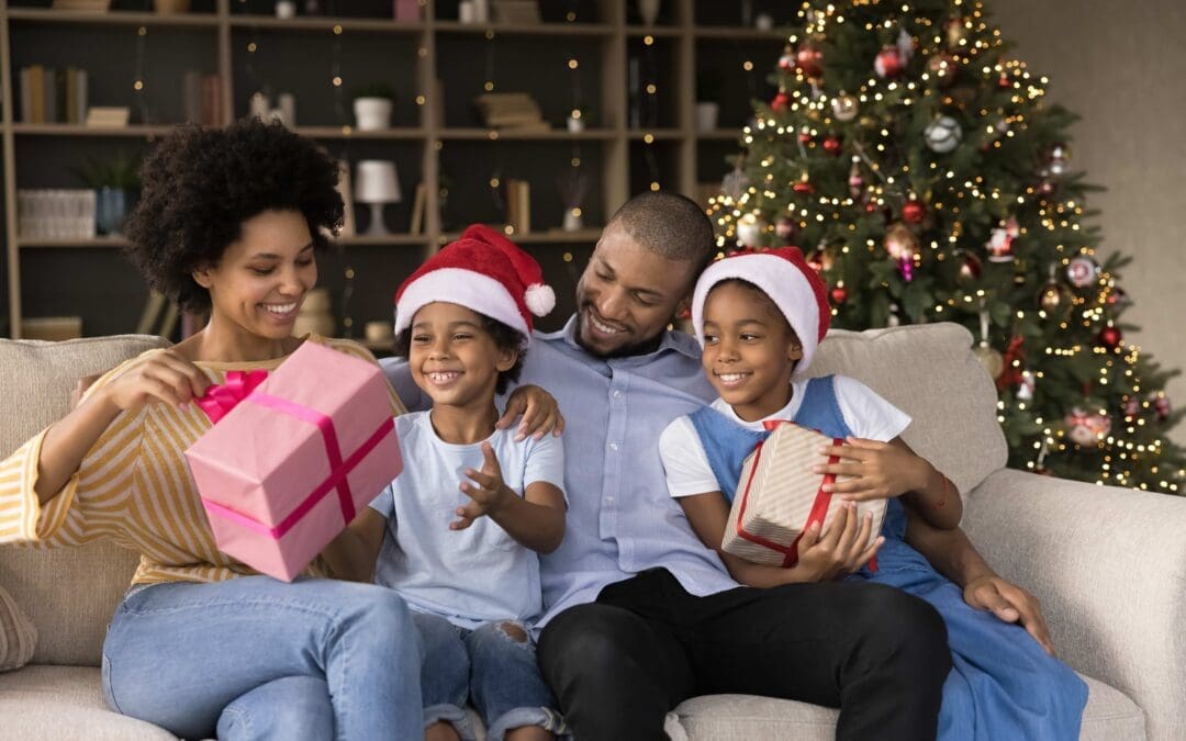 Smiling young african american parents having fun, unwrapping gift boxes with little children son daughter, resting on comfortable couch near decorated Christmas tree, winter holidays celebration.