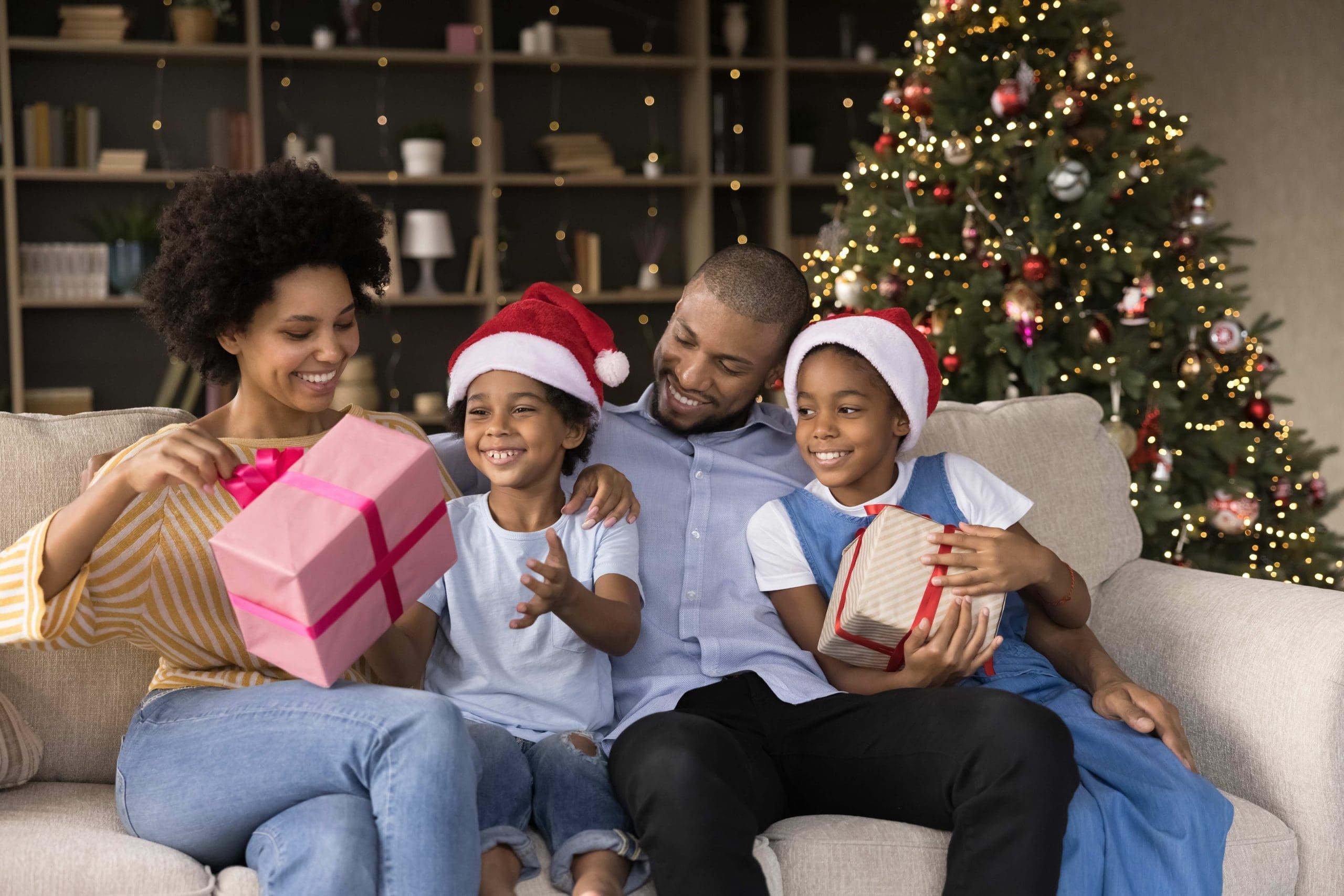 Smiling young african american parents having fun, unwrapping gift boxes with little children son daughter, resting on comfortable couch near decorated Christmas tree, winter holidays celebration.